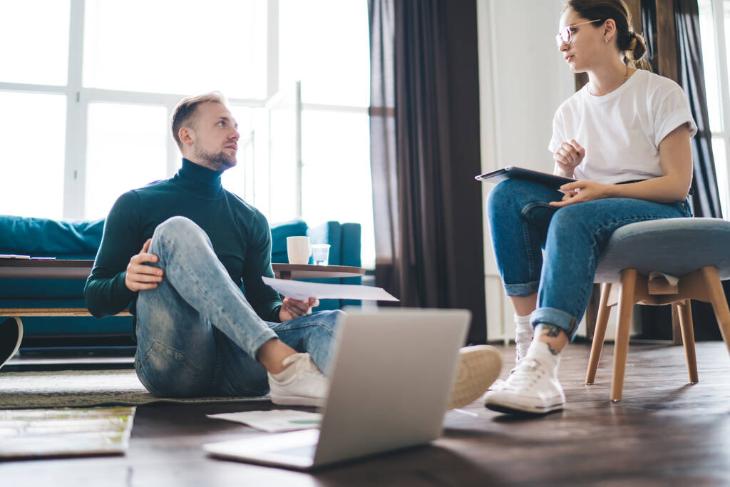 Low angle of young man and woman in casual clothes with laptop sharing thoughts about startup strategy while working together in modern workspace and looking at each other - Fotoğraf, Görsel