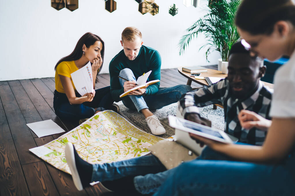 Group of concentrated multiracial coworkers in casual clothes sitting on floor in cozy office and gathering data while working on business project in team - Foto, imagen