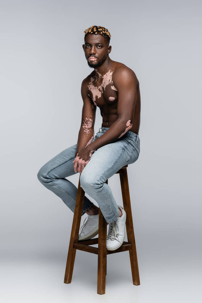 shirtless african american man with vitiligo looking at camera while sitting on stool in jeans on grey - Foto, Imagem