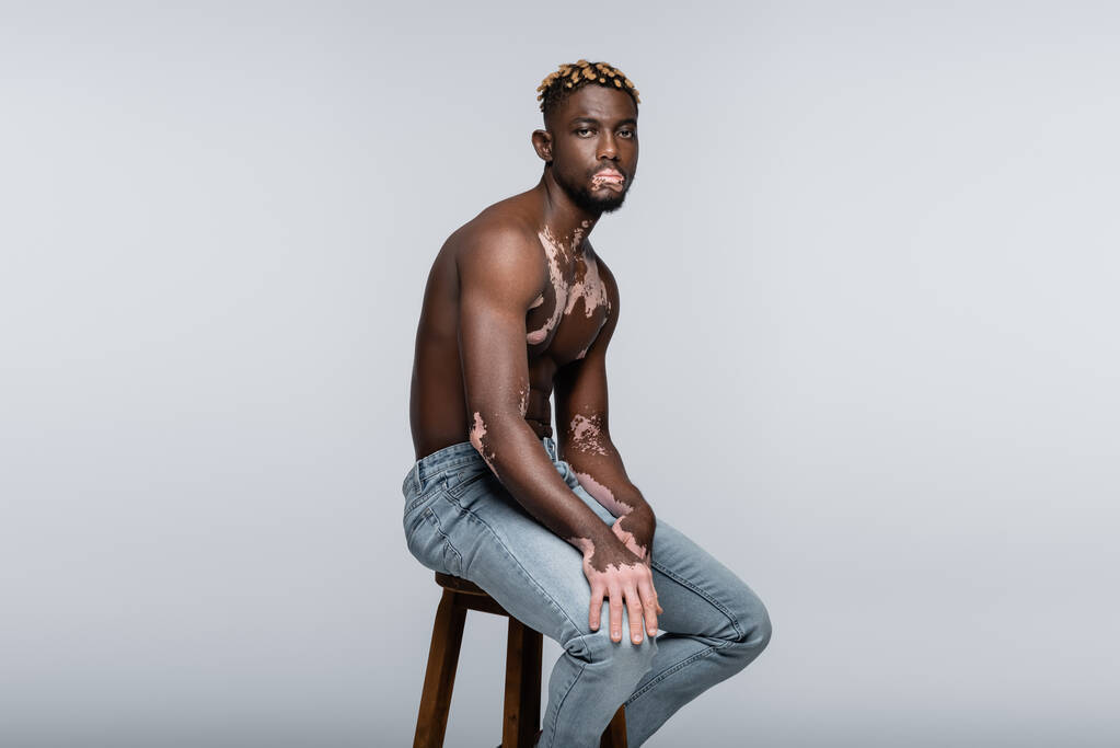 shirtless african american man with vitiligo looking at camera while sitting on stool in jeans isolated on grey - Foto, Imagem
