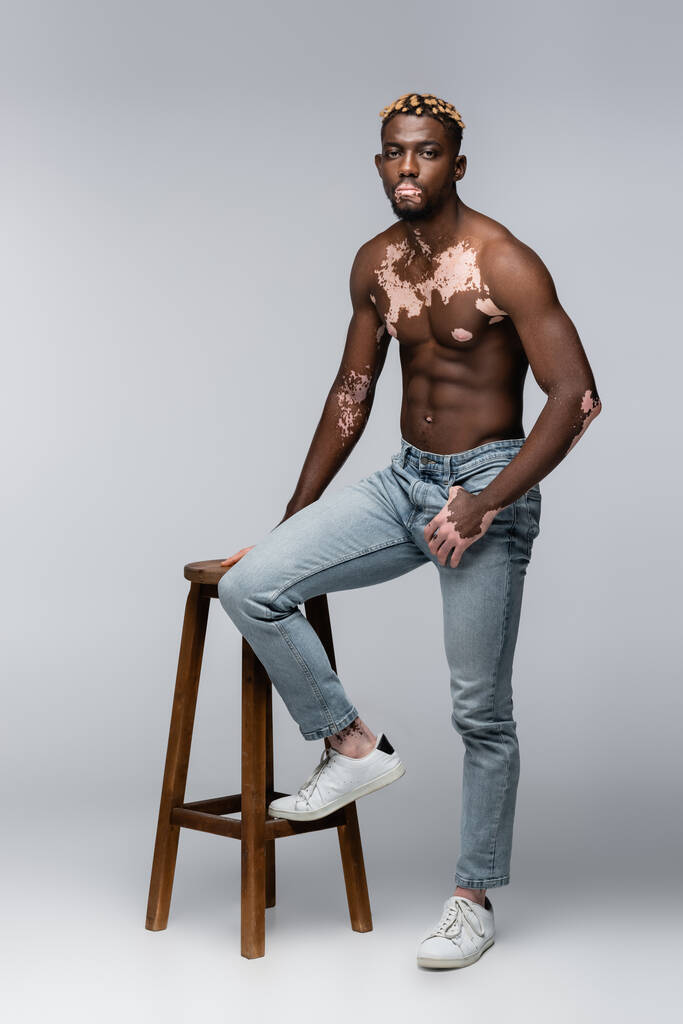 shirtless african american man with vitiligo and trendy hairstyle posing with thumb in pocket near high stool on grey - Zdjęcie, obraz