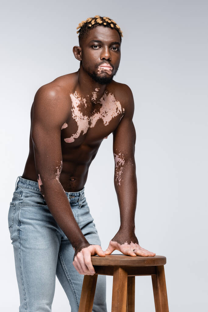 афроамериканський чоловік зі шкірою вітіліго і модною зачіскою, що стоїть без сорочки біля дерев'яного табурета на сірому
 - Фото, зображення
