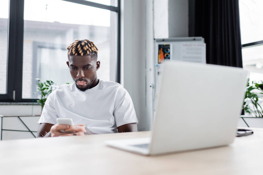 young african american man with vitiligo messaging on mobile phone near blurred laptop in office - Foto, imagen