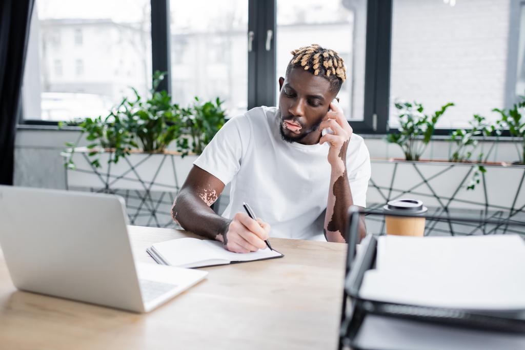 stylish african american man with vitiligo talking on smartphone and writing in notebook near blurred laptop - Foto, afbeelding
