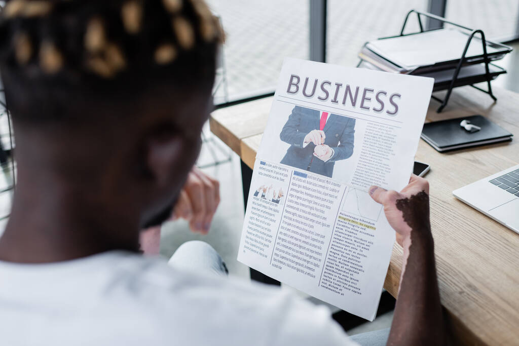 θολή αφροαμερικανός άνδρας με λεύκη ανάγνωση επιχειρηματική εφημερίδα κοντά σημειωματάριο και gadgets στο γραφείο - Φωτογραφία, εικόνα
