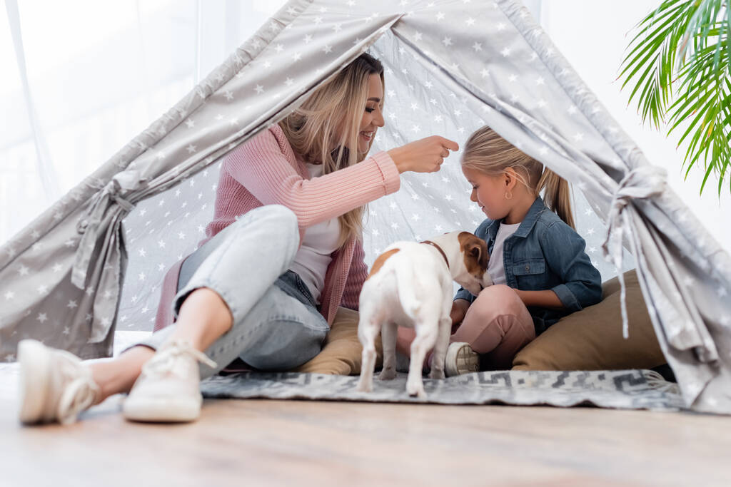 Positive mother and kid sitting in tent near jack russell terrier at home  - Fotografie, Obrázek