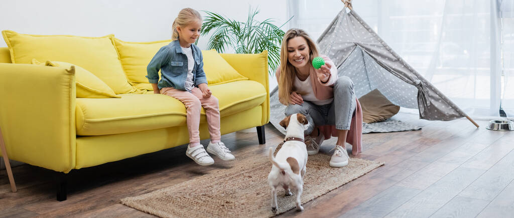 Positive woman holding toy near jack russell terrier and daughter on couch at home, banner  - Zdjęcie, obraz