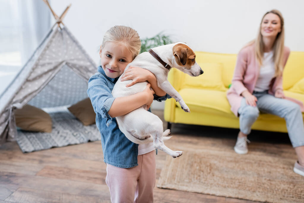 Happy kid hugging jack russell terrier near blurred mom at home  - Photo, Image