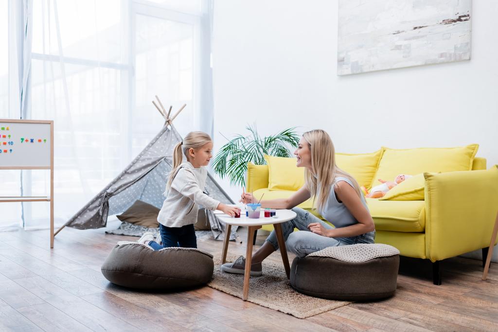 Side view of smiling mother and daughter drawing near couch and teepee at home  - Foto, Bild