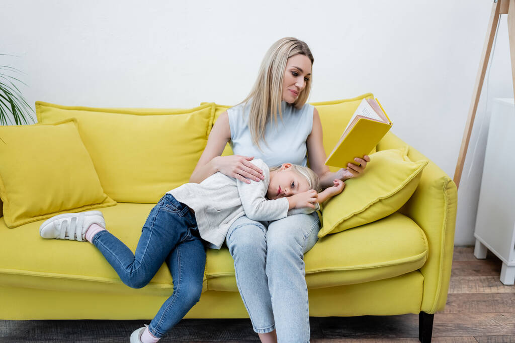 Мати читає книгу біля дитини на дивані вдома
  - Фото, зображення