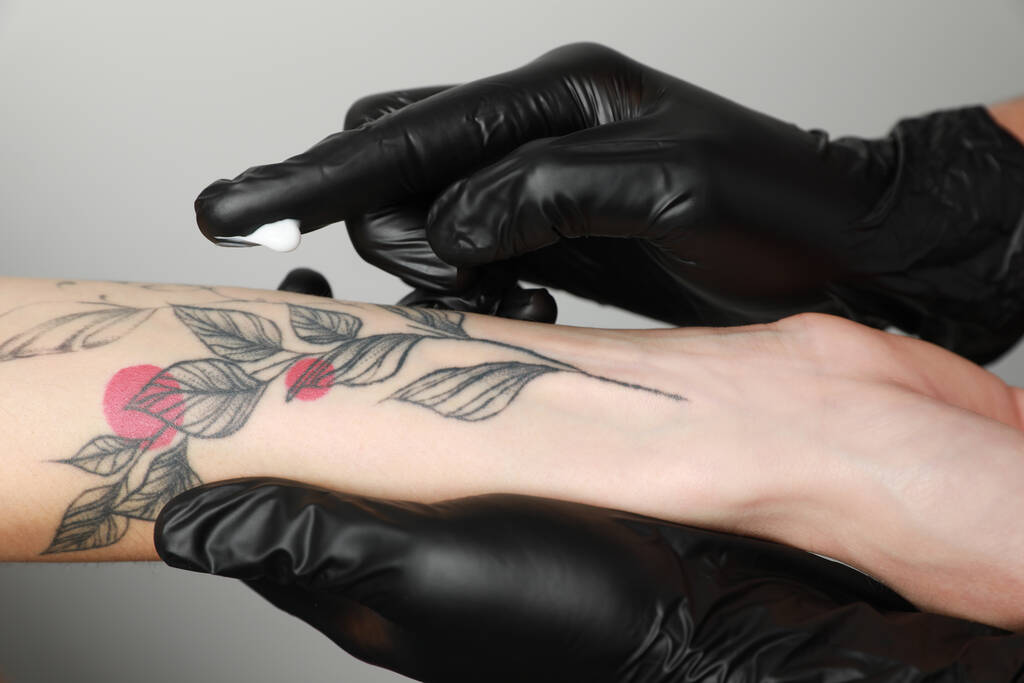 Worker in gloves applying cream on woman's arm with tattoo against light background, closeup - Fotó, kép