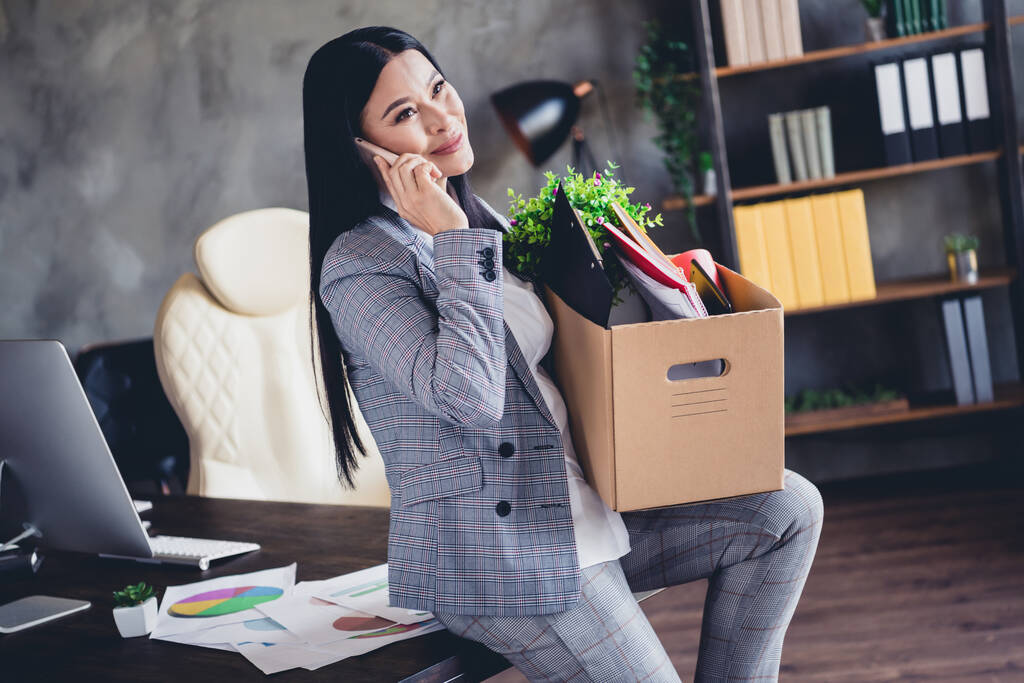 Фото привлекательной леди сидеть настольный пакет держать коробку говорить телефон носить серый пиджак офис внутри. - Фото, изображение