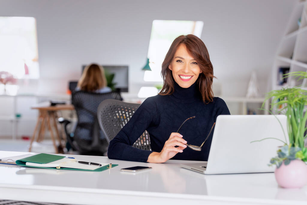 Pewna siebie bizneswoman w średnim wieku w swetrze turtleneck siedząc w biurze za laptopem i pracując nad nowym projektem.  - Zdjęcie, obraz