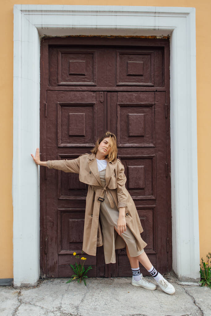 Jovem sorrindo mulher milenar com cabelo selvagem vestido com um casaco de outono posando perto da porta de um edifício antigo. - Foto, Imagem