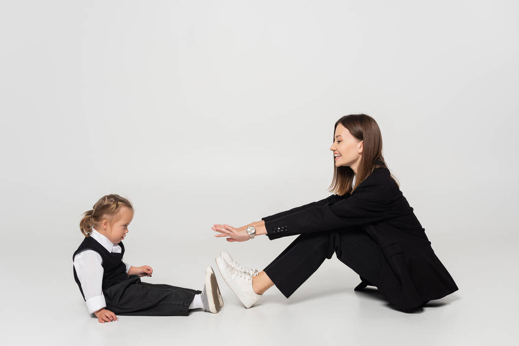 side view of cheerful mother sitting and reaching child with down syndrome on grey - Foto, afbeelding
