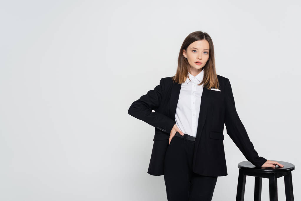young stylish woman in suit posing with hand on hip near chair isolated on grey - Photo, image