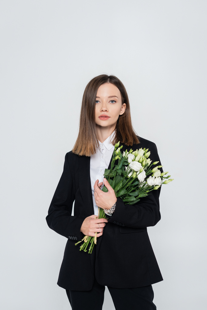 trendy young woman in black suit holding flowers isolated on grey  - Fotó, kép