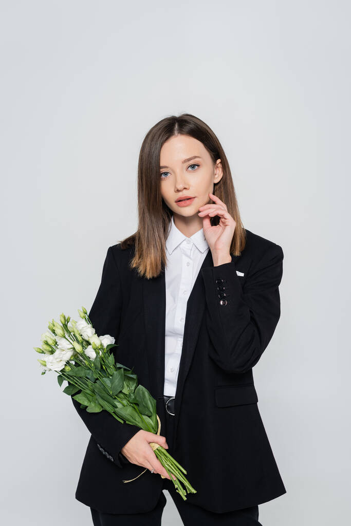 trendy woman in black suit holding flowers isolated on grey  - Foto, afbeelding