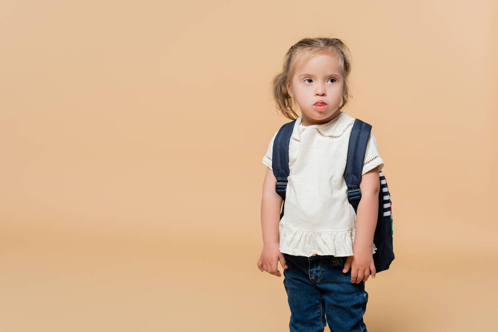 παιδί με σύνδρομο down προεξέχει γλώσσα ενώ στέκεται με σακίδιο στο μπεζ  - Φωτογραφία, εικόνα