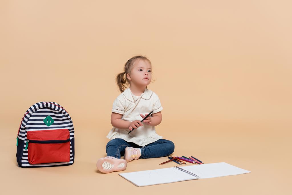 kid with down syndrome sitting near colorful pencils and backpack on beige - Foto, Bild