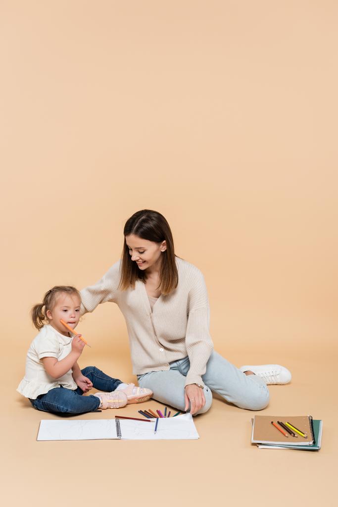 παιδί με σύνδρομο down κάθεται με ευτυχισμένη μητέρα κοντά πολύχρωμα μολύβια σε μπεζ - Φωτογραφία, εικόνα