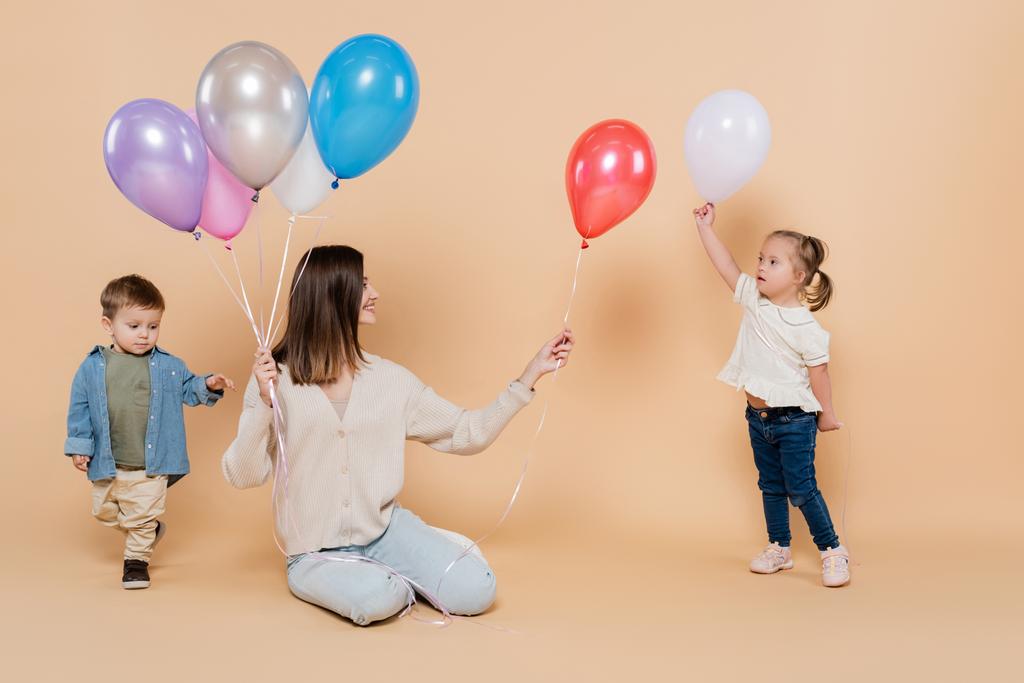 positive woman sitting near girl with down syndrome and toddler boy while holding colorful balloons on beige - Foto, Bild