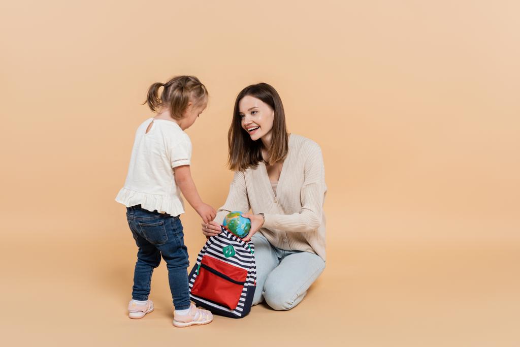 щаслива жінка тримає маленький глобус і рюкзак, дивлячись на дитину з синдромом Дауна на бежевому
 - Фото, зображення
