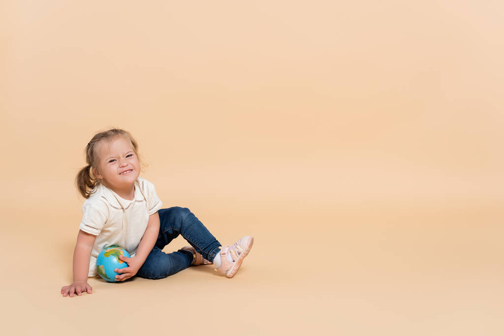 happy kid with down syndrome sitting with small globe on beige - Fotoğraf, Görsel