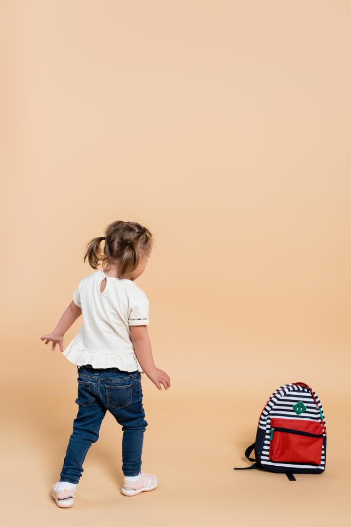 ダウン症候群の子供はベージュのバックパックの近くを歩く - 写真・画像