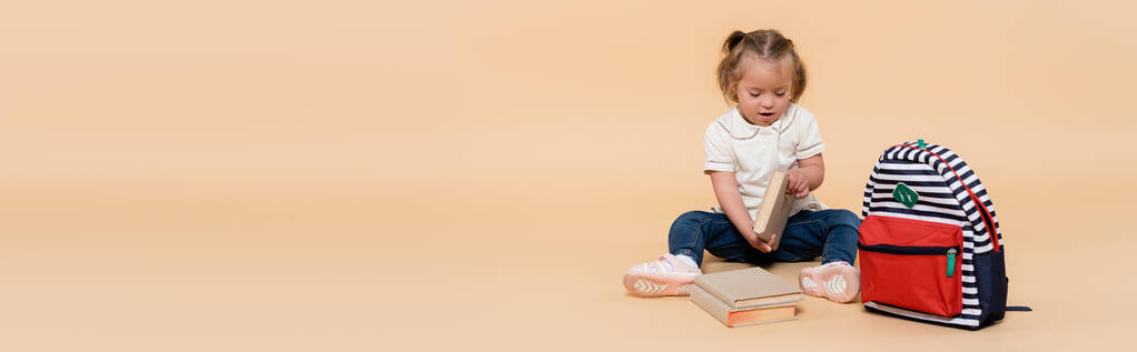 kid with down syndrome sitting near books and backpack on beige, banner - Foto, afbeelding