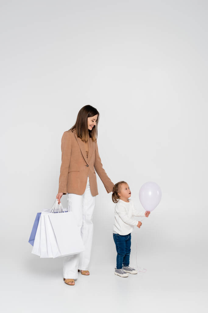 happy mother holding shopping bags and looking at girl with down syndrome near balloon on grey - 写真・画像