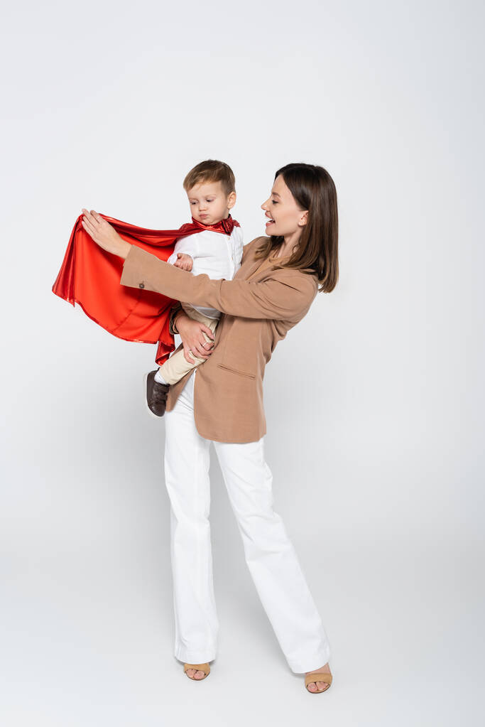 excited mother holding in arms toddler son in superhero costume on grey - Φωτογραφία, εικόνα