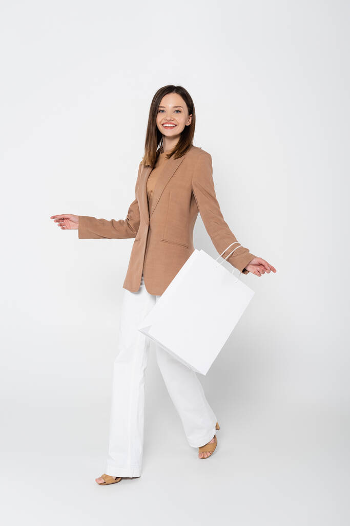 full length of joyful young woman in beige blazer holding shopping bag while posing on white - Fotó, kép