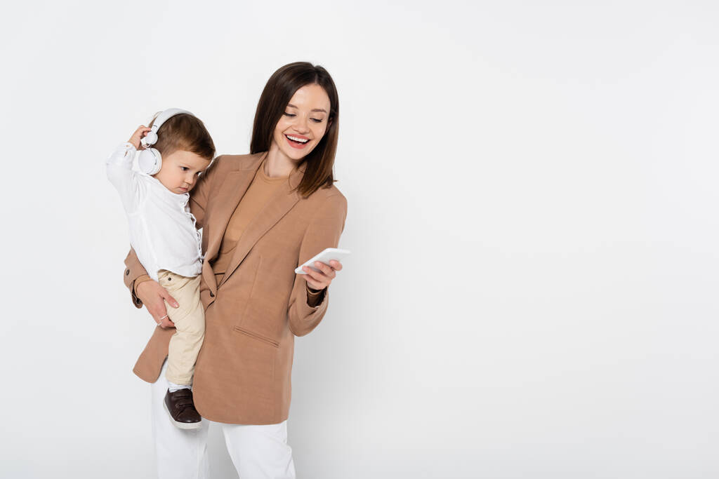 happy woman in beige blazer holding smartphone and toddler boy in headphones isolated on grey - Foto, Bild