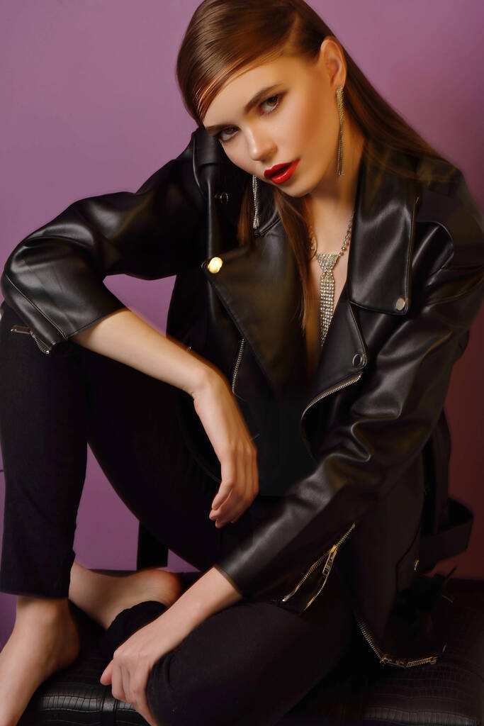 A young beautiful girl posing in a black leather jacket and gold jewelry on a background in a photo studio. - Photo, Image