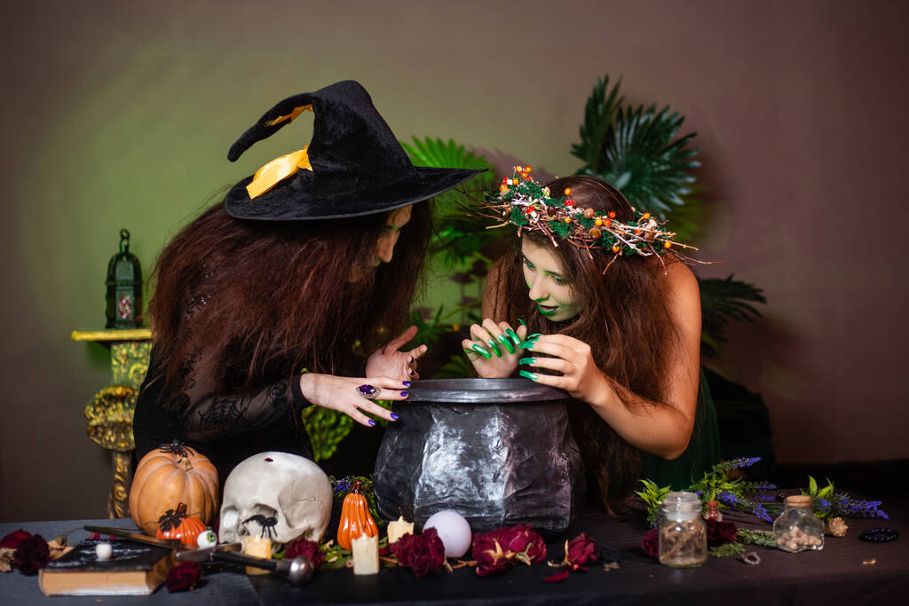 Two witches with disheveled hair brew a magic potion in a black cauldron, standing in a dark room. The concept of witchcraft on the eve of Saints' Day. - Foto, Bild