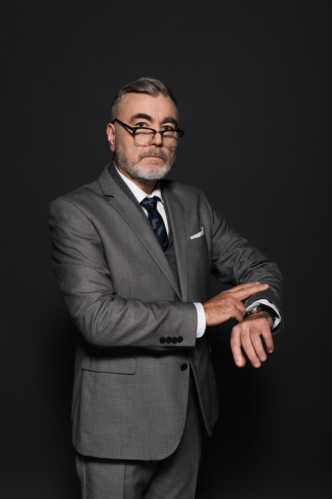 senior businessman in blazer, eyeglasses and wristwatch looking at camera on dark grey - Zdjęcie, obraz