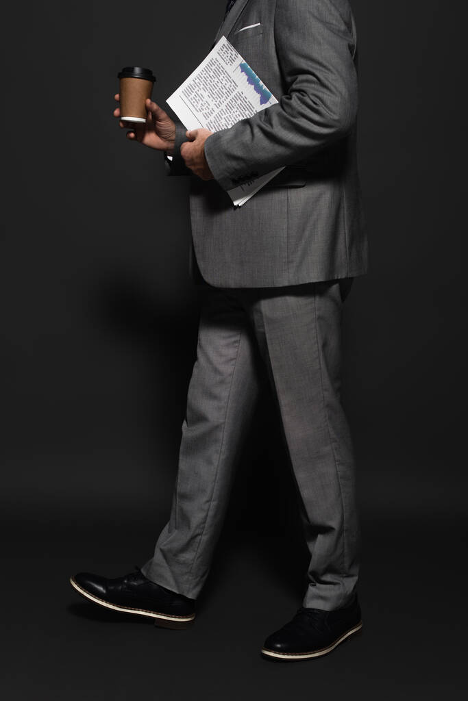 partial view of businessman in suit walking with newspaper and takeaway drink on dark grey - Fotografie, Obrázek