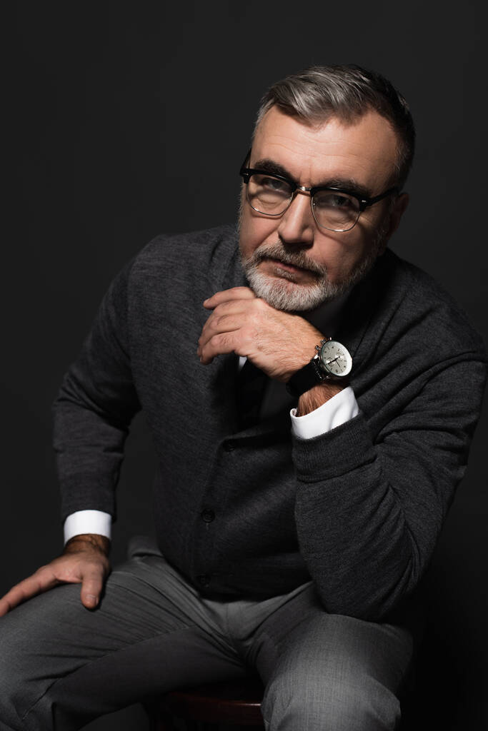 serious senior man in jumper, eyeglasses and wristwatch looking at camera while sitting on dark grey - Foto, Bild