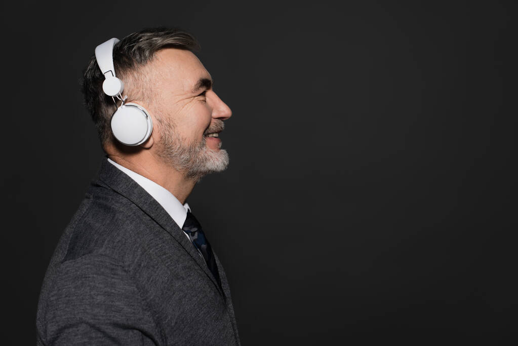 πλαϊνή άποψη του χαρούμενα γενειοφόρος άνθρωπος ακούγοντας μουσική σε ακουστικά που απομονώνονται σε μαύρο - Φωτογραφία, εικόνα