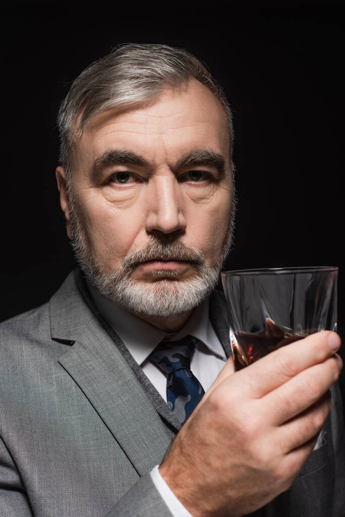 ανώτερος γενειοφόρος άνδρας σε σακάκι και γραβάτα κρατώντας ένα ποτήρι ουίσκι απομονωμένο σε μαύρο - Φωτογραφία, εικόνα