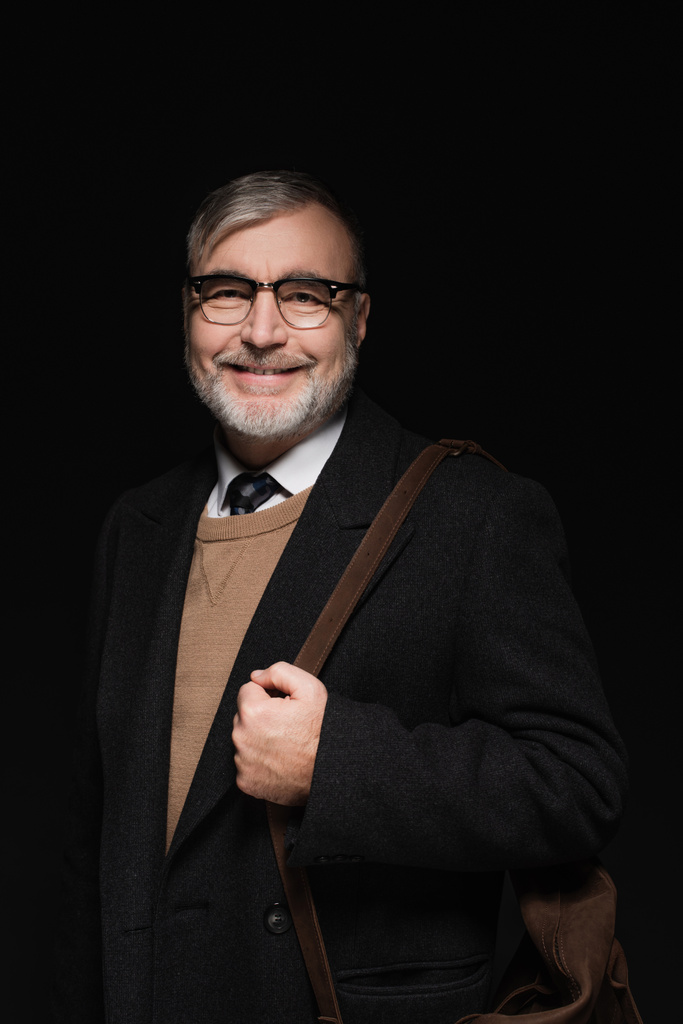 happy senior man in trendy coat and eyeglasses looking at camera isolated on black - Foto, immagini