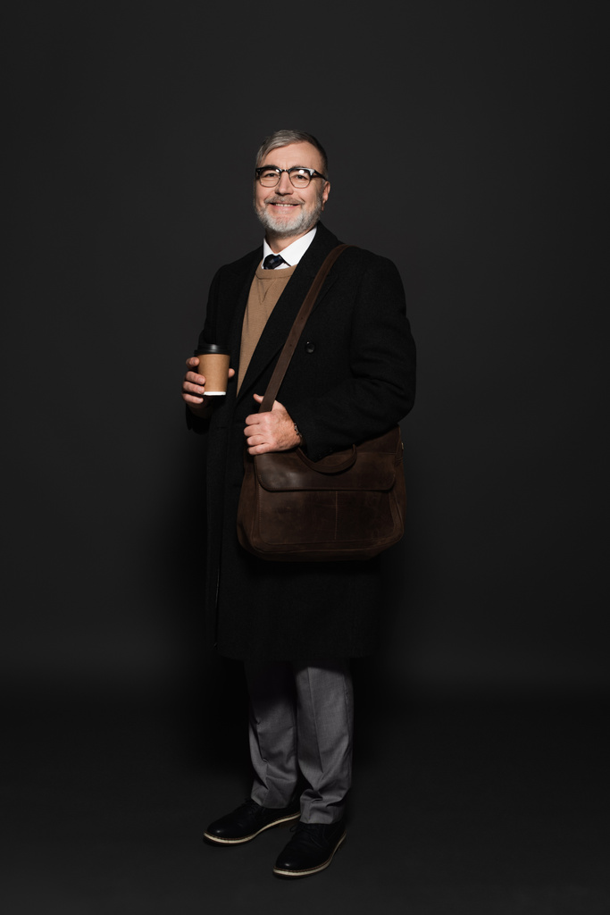 full length view of pleased senior man in black coat standing with leather bag on dark grey - Fotó, kép