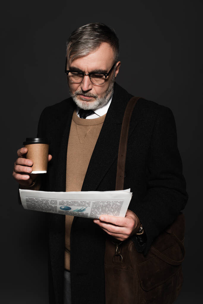 trendy senior man with leather bag and paper cup reading newspaper isolated on dark grey - Foto, afbeelding