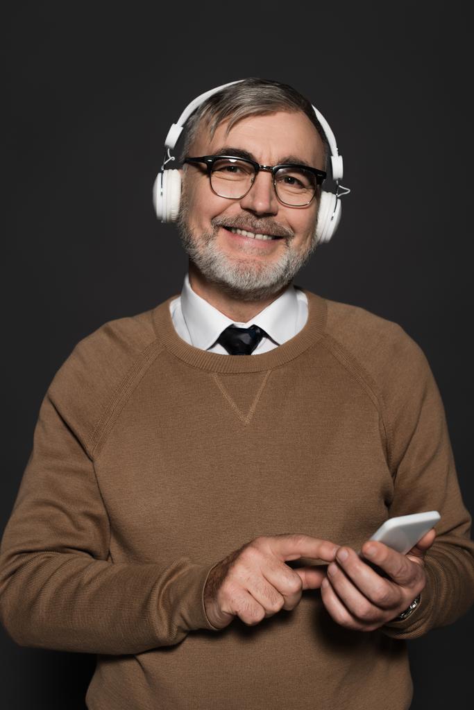 cheerful senior man in headphones looking at camera while holding smartphone isolated on black - Foto, imagen