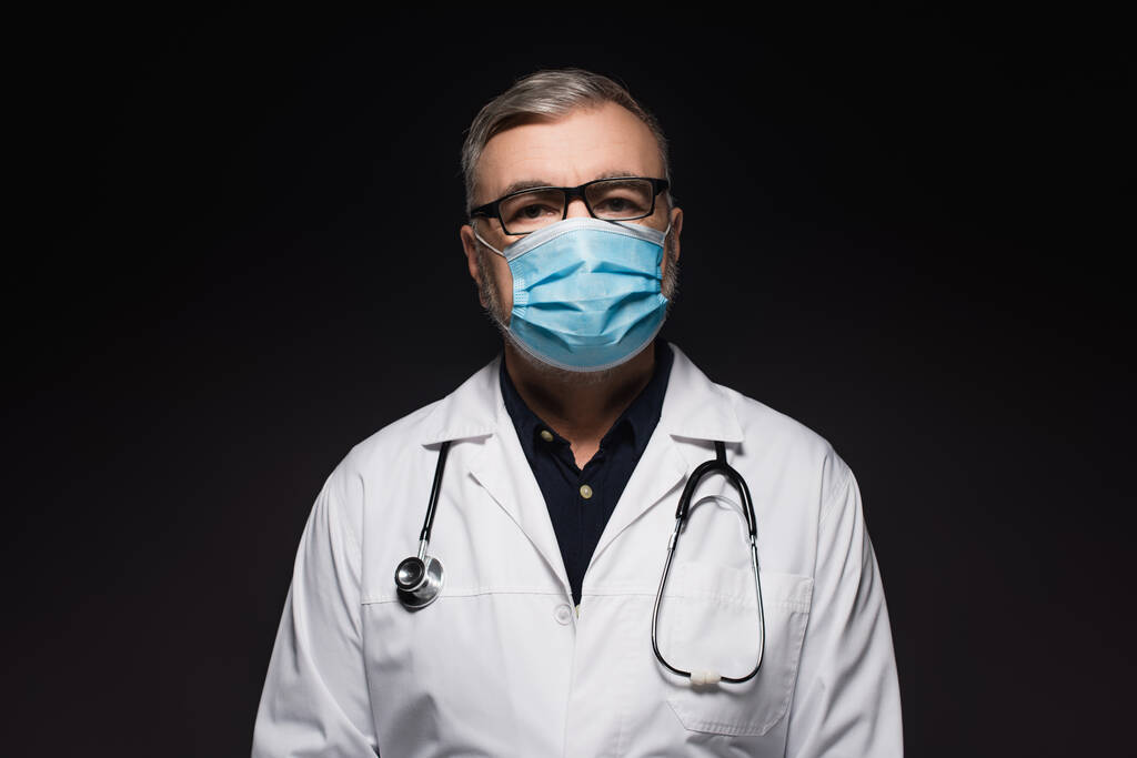старший лікар у білому пальто та медична маска, що дивиться на камеру ізольовано на чорному
 - Фото, зображення