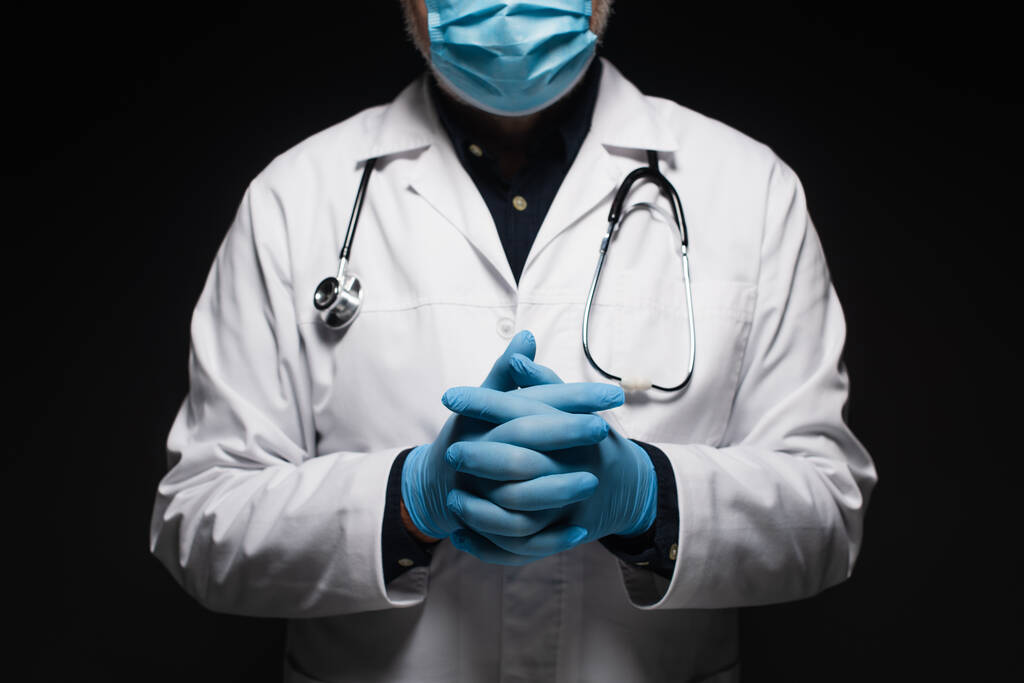 cropped view of doctor in medical mask and latex gloves standing with clasped hands isolated on black - Foto, afbeelding
