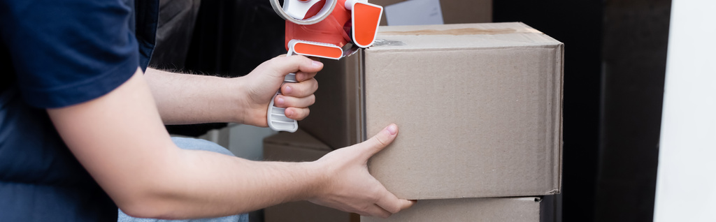 Cropped view of delivery man holding adhesive tape near cardboard boxes in car, banner  - Fotografie, Obrázek