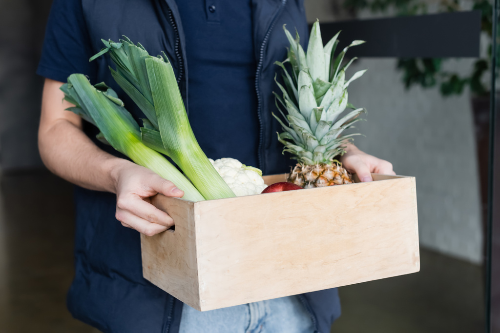 Cropped view of courier holding wooden box with fresh food in hallway  - Foto, Bild
