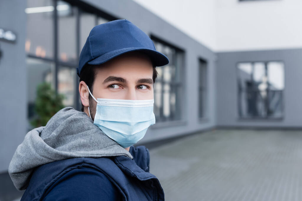Молодий чоловік доставки в медичній масці і кепці, дивлячись далеко на міську вулицю
  - Фото, зображення